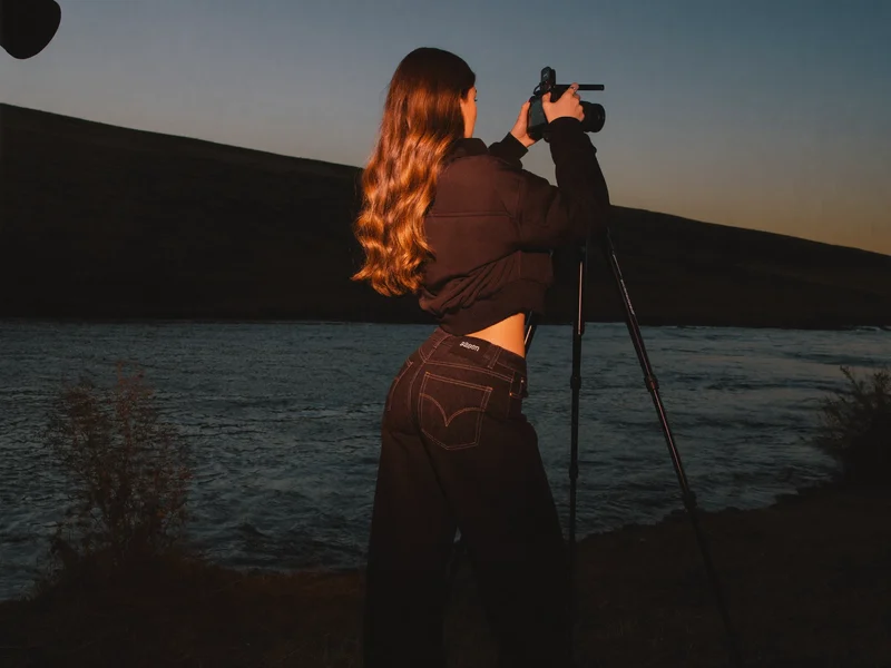 Fotografin von hinten mit Stativ an einem Flussufer in der Daemmerung